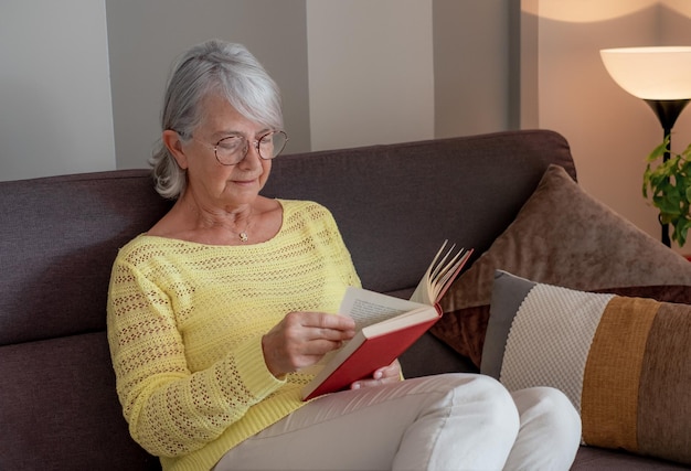 Retrato de uma mulher madura e relaxada, de cabelos grisalhos, deitada no sofá em casa, lendo um livro