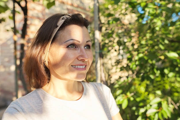 Foto retrato de uma mulher madura sorridente no jardim perto da casa, pôr do sol no rosto da mulher, copie o espaço