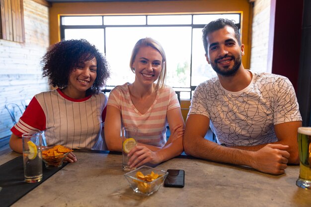 Retrato de uma mulher mestiça, uma mulher caucasiana e um homem mestiço sentados juntos no bar de um pub durante o dia, olhando para a câmera e sorrindo, com um smartphone, bebidas e lanches no b