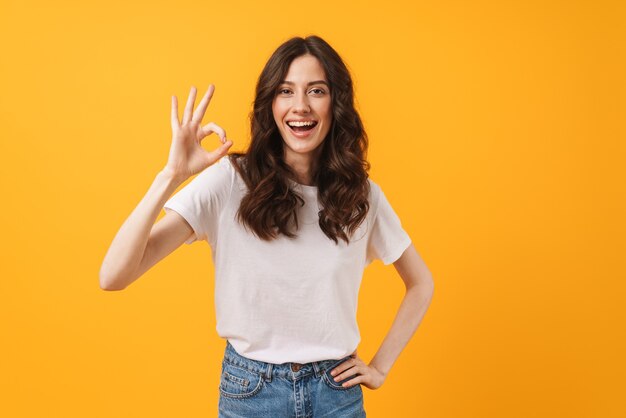 Retrato de uma mulher morena maravilhada, vestindo roupas casuais, sorrindo e mostrando sinal de ok para a câmera isolada sobre a parede amarela