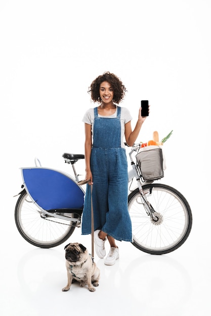 Retrato de uma mulher morena segurando um smartphone em pé com seu pug e uma bicicleta isolada sobre uma parede branca