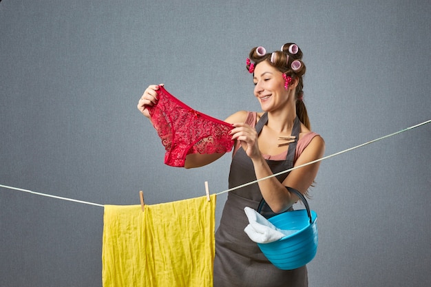 Retrato de uma mulher muito retrô pendurando a calcinha em uma corda na parede cinza. Fanny dona de casa vestindo rolos secando roupas. Conceito doméstico.