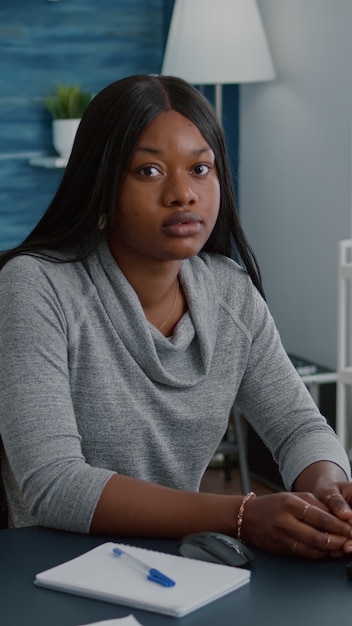 Retrato de uma mulher negra alegre olhando para a câmera enquanto está sentado na mesa da escrivaninha na sala de estar durante ...