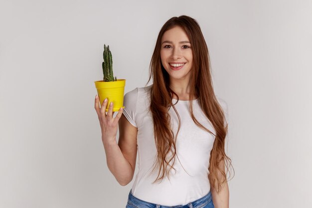 Retrato de uma mulher positiva satisfeita segurando o vaso de flores amarelas e cactos olhando para a câmera com sorriso de dentes vestindo camiseta branca Foto de estúdio interior isolada em fundo cinza