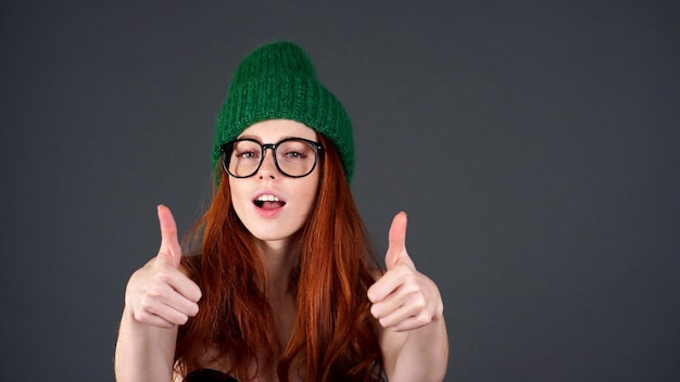 Retrato de uma mulher ruiva com um chapéu verde com um sorriso largo aparece os polegares com as duas mãos
