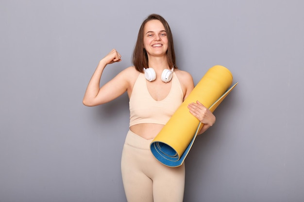 Retrato de uma mulher satisfeita e encantada com cabelo castanho em pé tapete de ioga e fones de ouvido no pescoço mostrando seu bíceps sorrindo aproveitando o treino e o resultado da perda de peso