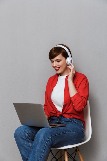 Retrato de uma mulher satisfeita em fones de ouvido, sorrindo enquanto está sentado na cadeira com um laptop isolado na parede cinza