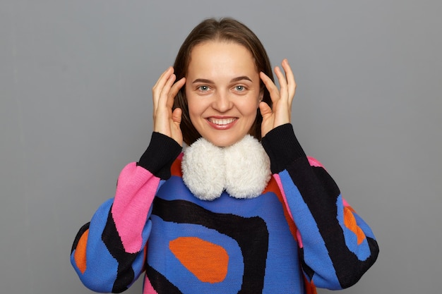 Foto retrato de uma mulher satisfeita sorridente e encantada com cabelos castanhos vestindo um jumper colorido posando isolado sobre um fundo cinza tocando seu cabelo olhando para a câmera expressando felicidade