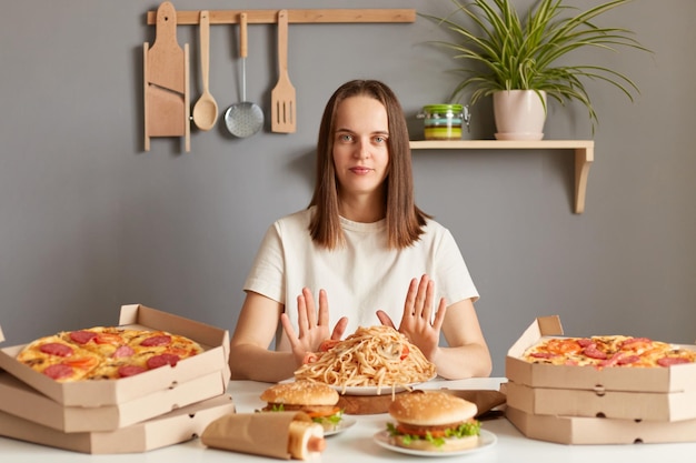 Retrato de uma mulher séria e confiante com cabelos castanhos vestindo camiseta casual branca sentada à mesa na cozinha mantém a dieta mostrando o gesto de parada não come junk food prefere uma alimentação saudável