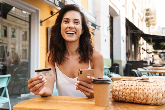 Retrato de uma mulher simpática e encantada com um saco de palha segurando um cartão de crédito e usando o celular enquanto está sentado em um café aconchegante ao ar livre