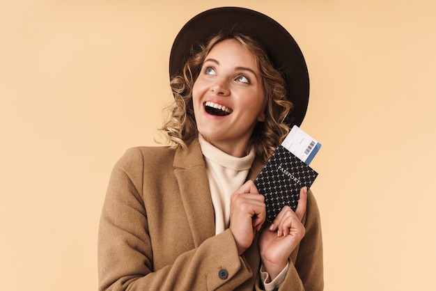 Foto retrato de uma mulher sorridente a sonhar com chapéu isolado na parede bege, segurando o passaporte com ingressos, olhando de lado.