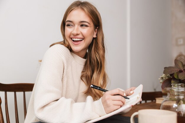 Retrato de uma mulher sorridente caucasiana, vestindo roupas casuais, fazendo anotações e bebendo chá em um quarto aconchegante