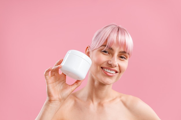 Retrato de uma mulher sorridente com cabelo rosa segurando um frasco branco com loção hidratante corporal após