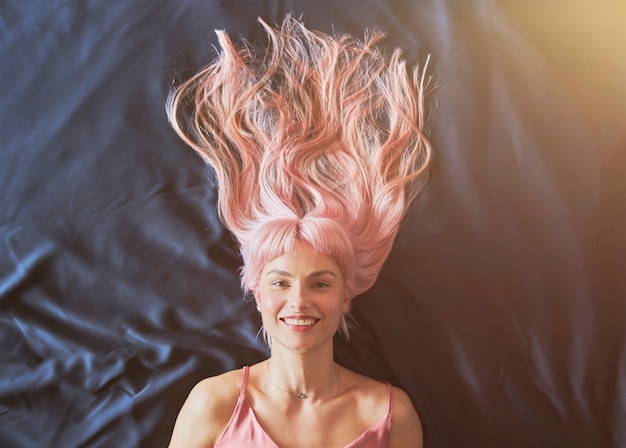 Retrato de uma mulher sorridente com cabelo rosa solto, deitada em uma cama grande com lençol azul escuro