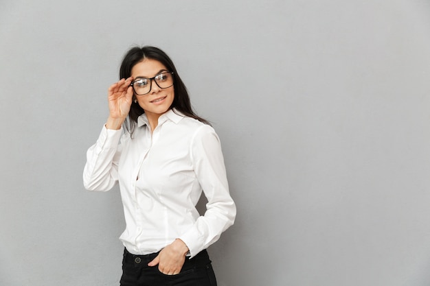 Retrato de uma mulher sorridente de sucesso com longos cabelos castanhos, vestindo roupas de escritório, olhando para o lado e tocando seus óculos, isolado sobre um fundo cinza