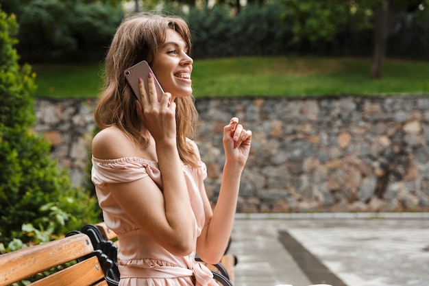 Retrato de uma mulher sorridente e animada usando um vestido, falando no celular, enquanto está sentada no banco de um parque