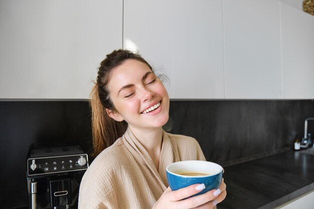 Retrato de uma mulher sorridente e despreocupada tomando café em pé na cozinha com um rosto encantado e satisfeito