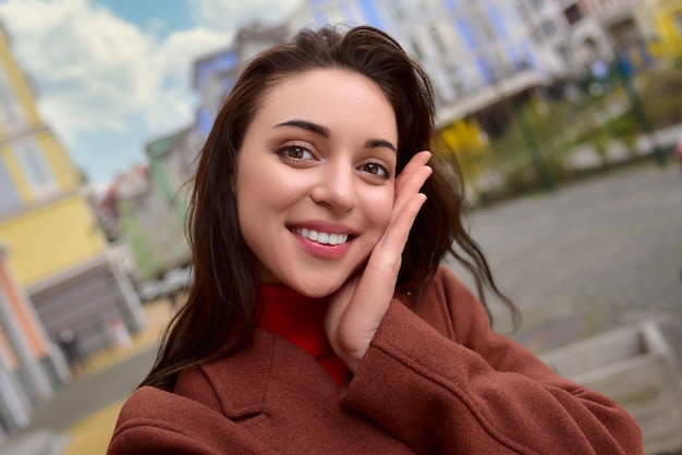 Retrato de uma mulher sorridente e elegante com um casaco castanho de outono tira uma selfie na rua