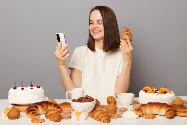 Retrato de uma mulher sorridente e encantada com cabelos castanhos, sentada à mesa com sobremesas, segurando um telefone inteligente, fazendo uma chamada de vídeo ou fazendo uma selfie com um croissant na mão isolado sobre fundo cinza