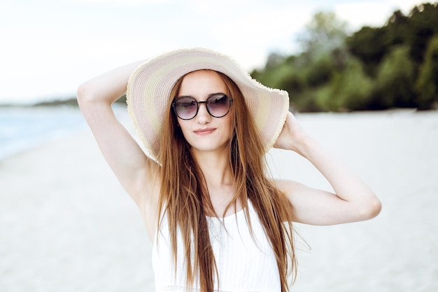 Retrato de uma mulher sorridente feliz em êxtase de felicidade livre na praia do oceano em pé com um chapéu e óculos de sol. Uma modelo feminina em um vestido branco de verão curtindo a natureza durante as férias de viagem