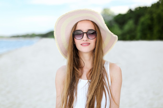 Retrato de uma mulher sorridente feliz em êxtase de felicidade livre na praia do oceano em pé com um chapéu e óculos de sol. Uma modelo feminina em um vestido branco de verão curtindo a natureza durante as férias de viagem