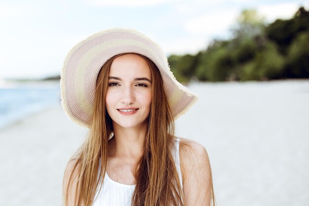 Retrato de uma mulher sorridente feliz em êxtase de felicidade livre na praia do oceano em pé com um chapéu. Um modelo feminino em um vestido branco de verão curtindo a natureza durante as férias de viagem ao ar livre