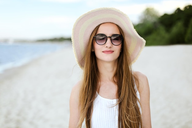Retrato de uma mulher sorridente feliz em êxtase de felicidade livre na praia oceânica em pé com um chapéu e óculos de sol. Uma modelo feminina em um vestido branco de verão curtindo a natureza durante as férias de viagem