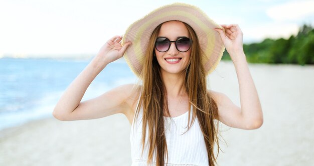 Retrato de uma mulher sorridente feliz em felicidade livre na praia do oceano de pé com um chapéu e óculos de sol. Uma modelo feminina em um vestido de verão branco desfrutando da natureza durante as férias de viagem