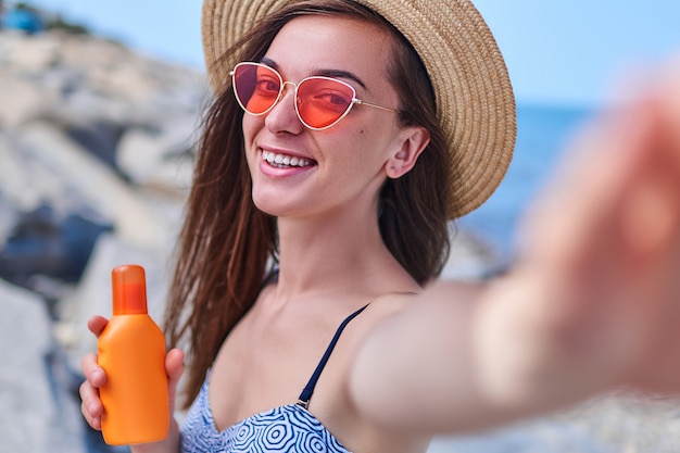 Retrato de uma mulher sorridente feliz em um maiô, chapéu de palha e óculos de sol vermelhos brilhantes com uma garrafa de protetor solar durante o banho de sol à beira-mar no mar em tempo ensolarado no verão