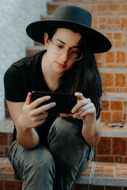 Retrato de uma mulher usando o telefone celular, usando óculos um chapéu preto.