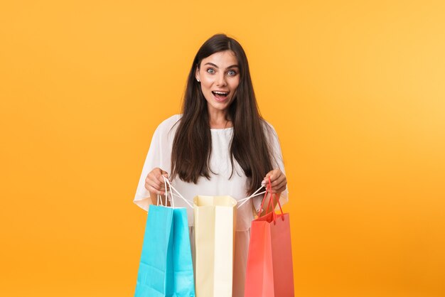 Retrato de uma mulher viciada em compras usando um vestido e sorrindo enquanto segura sacolas de compras coloridas isoladas na parede amarela