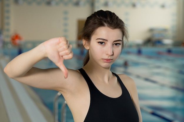 Retrato de uma nadadora em forma gesticulando com os polegares para baixo à beira da piscina no centro de lazer.
