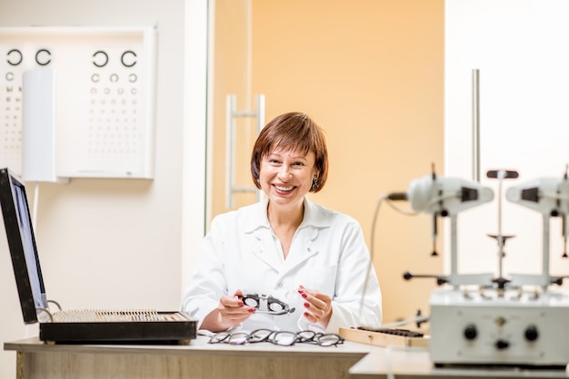 Retrato de uma oftalmologista sênior em uniforme sentada com diferentes dispositivos ópticos e óculos durante o trabalho no escritório