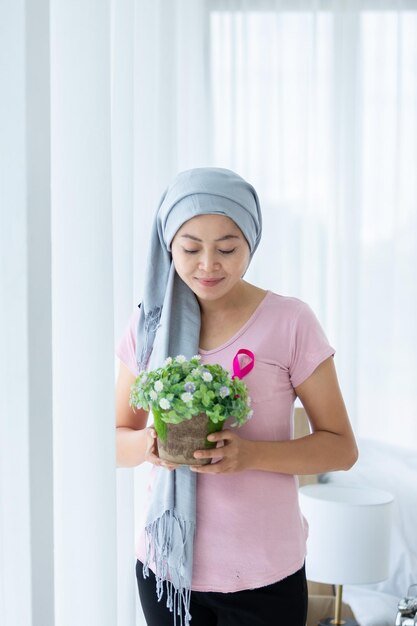 Retrato de uma paciente com câncer mamário com doença de mulher asiática Segurando vasos de flores com fita rosa usando lenço na cabeça Após o tratamento de quimioterapia na janela No quarto da casa