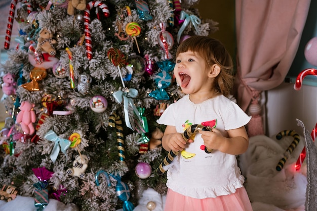 Retrato de uma pequena menina loira brincando perto da árvore de Natal, a criança está vestindo um ts branco.