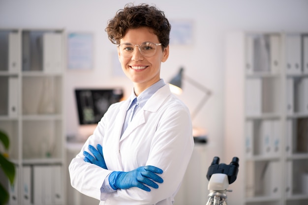 Retrato de uma pesquisadora de laboratório, mulher confiante e positiva, de óculos e jaleco branco, de braços cruzados