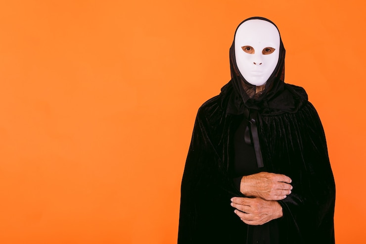Foto retrato de uma pessoa com máscara assassina branca de halloween, capa e capuz, olhando para a câmera, vestida para o halloween contra um fundo laranja, com espaço de cópia à esquerda