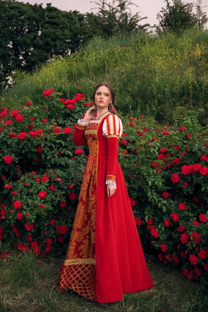 Foto retrato de uma princesa medieval de fantasia em um vestido vermelho vintage mulher em pé no jardim de rosas