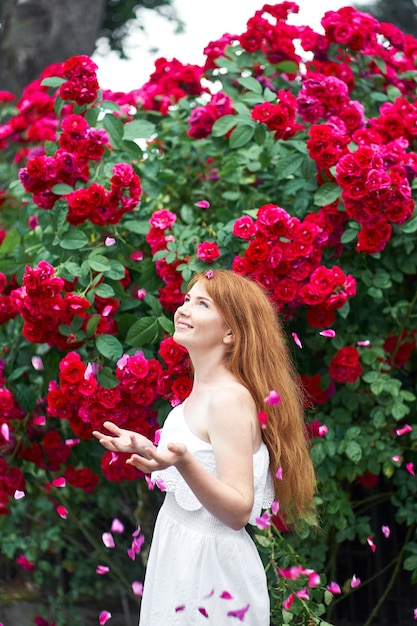 Retrato de uma ruiva bonita, vestida com um vestido de luz branca sobre um fundo de rosas florescendo