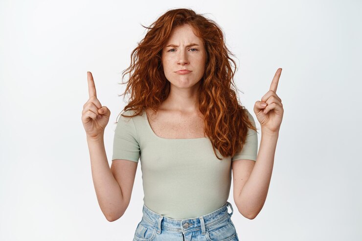 Foto retrato de uma ruiva mal-humorada apontando os dedos para cima em algo injusto sendo decepcionado em pé sobre fundo branco copiar espaço