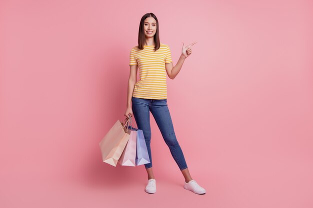 Retrato de uma senhora alegre e confiável segurando sacolas de loja direta com o dedo no espaço vazio no fundo rosa