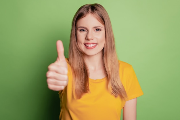 Retrato de uma senhora de confiança do promotor demonstra o polegar para cima vestindo uma camiseta amarela isolada no fundo de cor verde