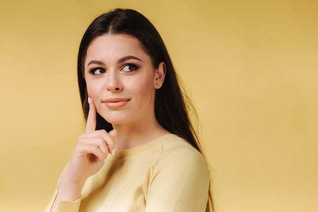 Retrato de uma senhora incrível com maquiagem pensando com braço no queixo e suéter amarelo casual