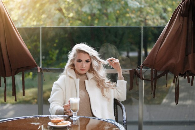 Retrato de uma senhora loira em uma mesa em um café, cabelo comprido, casaco bege. Posando para a câmera.