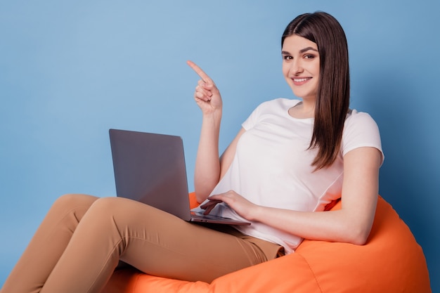 Retrato de uma senhora relaxada sentar com o pufe segurando o espaço vazio do dedo direto do netbook no fundo azul