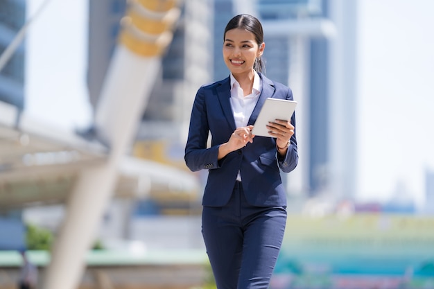 Retrato de uma sorridente mulher de negócios segurando um tablet digital e caminhando em frente a modernos edifícios de escritórios