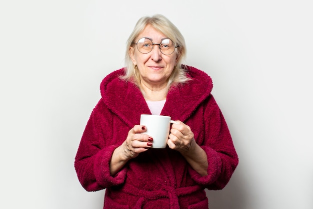 Retrato de uma velha amigável em um roupão casual e óculos, segurando um copo em uma parede leve isolada. Rosto emocional. Conceito de café da manhã