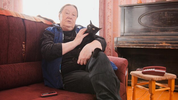 Retrato de uma velha em casa - senhora sentada no sofá com um gato preto, telefoto