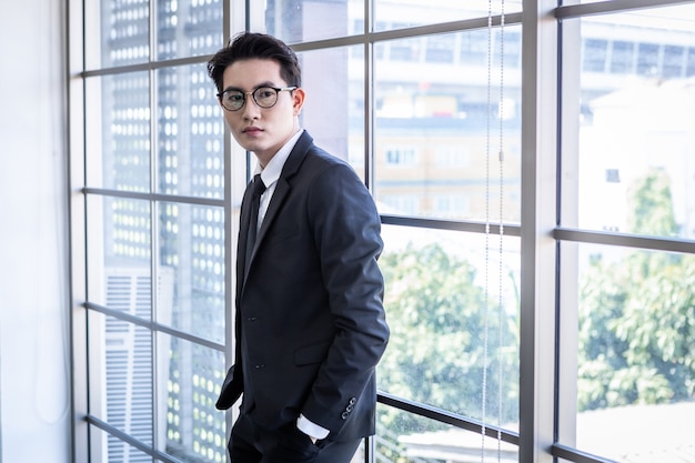 Retrato de vencedor de sentimento bem sucedido bonito jovem empresário asiático com óculos usa um terno de homem de jaqueta azul e camisa azul, olhando para a janela no fundo da sala de escritório.