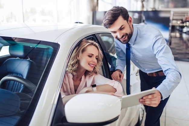 Foto retrato de vendedor sorridente falando com cliente do sexo feminino sentado no carro usando tablet digital no salão de concessionária